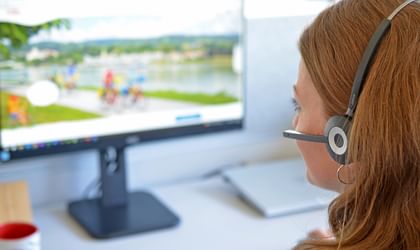 Vrouw met lang blond haar draagt een headset en zit aan een wit bureau met computermonitor, toetsenbord en koffiemok.