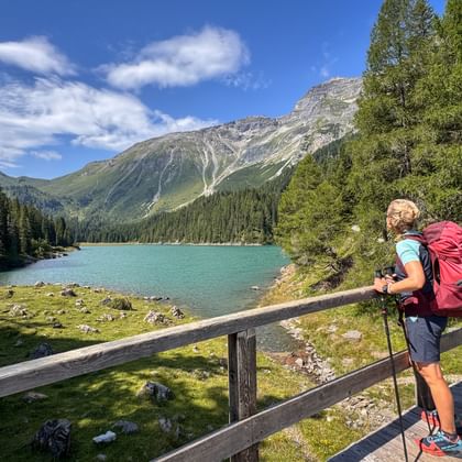 Wandelaar met rode rugzak en wandelstokken staat op houten brug met uitzicht op turquoise Obernberger See omringd door bergen.