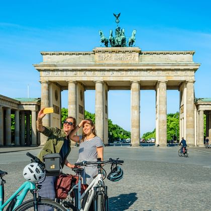 Twee vrouwelijke fietsers nemen een selfie voor de Brandenburger Tor in Berlijn. Ze staan met hun fietsen op het geplaveide plein.