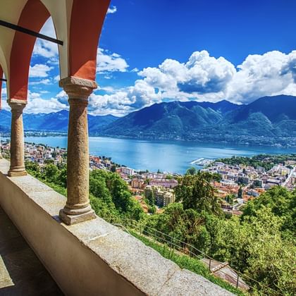 Panoramisch uitzicht vanaf Santa Caterina kloosterterras over Lago Maggiore met bergen, stadsgebouwen en blauwe lucht met wolken.