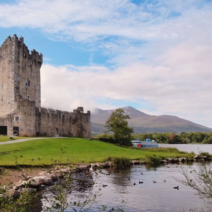 Ross Castle, een middeleeuwse stenen vesting met hoge toren, staat op groene gronden naast een meer in Killarney, Ierland, met bergen op de achtergrond.
