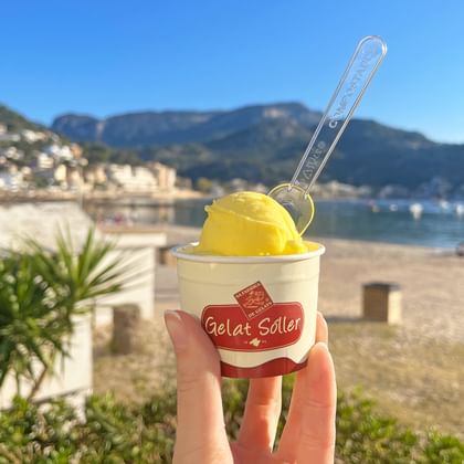 Hand houdt sinaasappelijs-bekertje met lepel vast tegen achtergrond van Port de Sóller strand met baai, bergen en palmbomen.