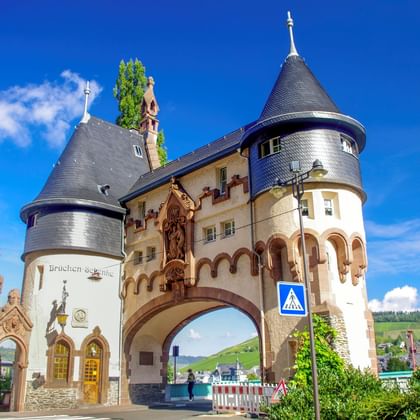 Historische brugpoort met twee ronde torens en kegelvormige daken in Traben-Trarbach onder blauwe hemel. Wijngaarden zichtbaar op heuvels op achtergrond.
