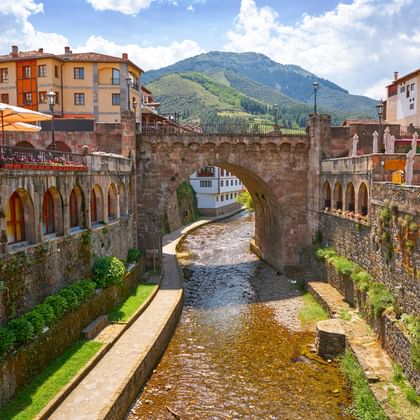 Stenen brug over rivier in Potes, Cantabrië, met kleurrijke gebouwen en groene bergen onder blauwe lucht met witte wolken.