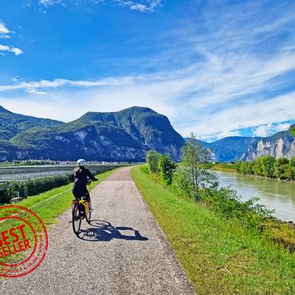 Fietser op pad in Dolomieten vallei Fietser rijdt op geasfalteerd pad door groene vallei in Dolomieten met dramatische bergtoppen, rivier en blauwe lucht met witte wolken.