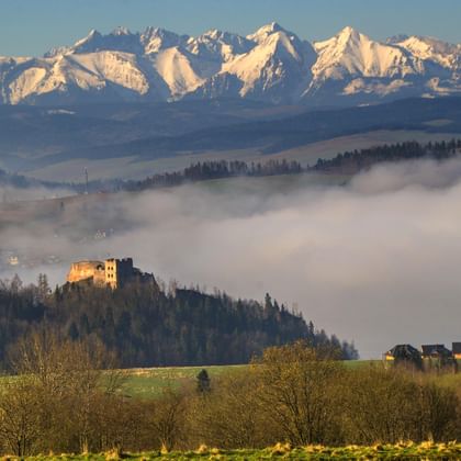 Besneeuwde Beskiden in Polen boven mistige valleien. Een kasteel op een beboste heuvel op de middenvlakte met groene weiden ervoor.