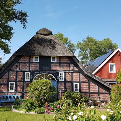 Traditionele Noord-Duitse boerderij met rieten dak en bakstenen muren, witte ramen en blauwe bank. Een rood houten huis staat erachter.