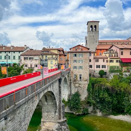 Stenen brug met rode afzetting over groene rivier in Cividale. Kleurrijke historische gebouwen en klokkentoren onder bewolkte hemel.