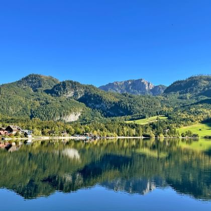 Perfecte spiegelreflectie van bergen en dorp in kalme Grundlsee wateren. Beboste Alpentoppen omringen het meer onder heldere blauwe lucht.