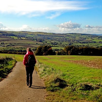 Wandelaar in rode jas loopt op landweg door Cotswolds landschap met groene heuvels en velden onder blauwe lucht.