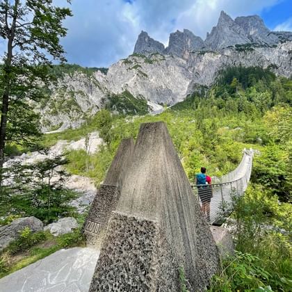 Hangbrug in Klausbach vallei, Nationaal Park Berchtesgaden, met wandelaars en dramatische Alpentoppen op de achtergrond.
