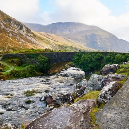 Mosbedekte stenen brug kruist een rotsachtige beek in Killarney, Ierland. Bergen rijzen op de achtergrond onder bewolkte luchten.
