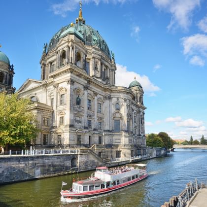 Berlijnse Dom met groene koperen koepels aan de Spree. Toeristische boten varen op het water terwijl bezoekers langs de oeverpromenade wandelen.