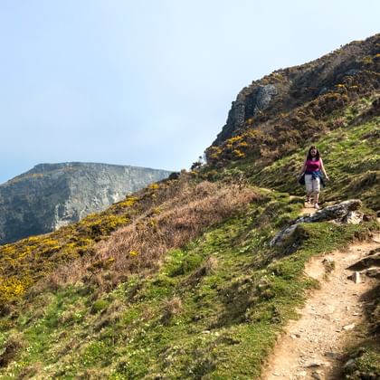Wandelaar op een kustpad in Cornwall met rotsachtige kliffen, groene vegetatie, gele bremstruiken en uitzicht op de blauwe oceaan.