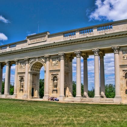 Neoklassieke colonnade met Korinthische zuilen en centrale boog in Valtice, Tsjechië. De beige stenen structuur staat op een gazon onder blauwe lucht.
