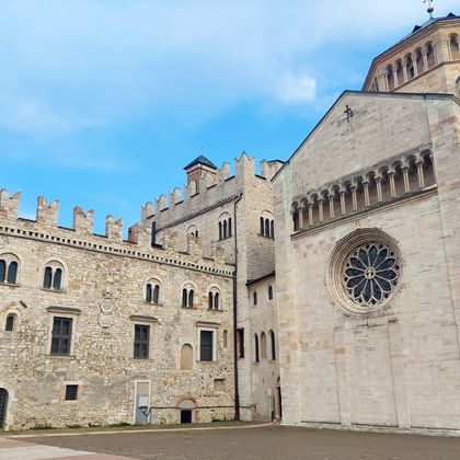 Kathedraal van Trento met roosvenster en klokkentoren naast middeleeuws paleis met kantelen onder blauwe hemel met witte wolken.