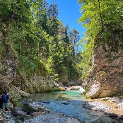 Wandelaar met rugzak bij turquoise rivier door rotsachtige kloof. Houten loopbrug zichtbaar op linkse rotswand, omringd door bos.