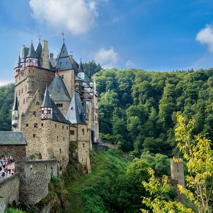 Burg Eltz, een middeleeuws kasteel met meerdere torens en torentjes, gelegen op een heuvel omringd door dicht groen bos onder een blauwe lucht.
