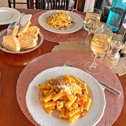 Twee pasta gerechten op witte borden met broodmand en witte wijnglazen op houten tafel. Eén bord heeft rigatoni met tomatensaus en kaas.