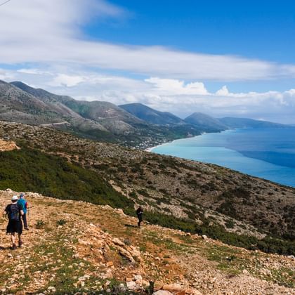 Twee wandelaars met rugzakken lopend op rotsachtig bergpad met uitzicht op de Albanese kust bij Borsh met blauwe zee en bergen.