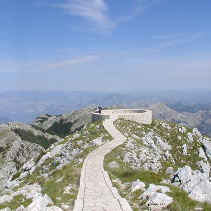 Stenen pad kronkelt over rotsachtige Lovćen-top naar rond uitkijkpunt, met bergketens en kustzicht onder blauwe lucht in Montenegro.