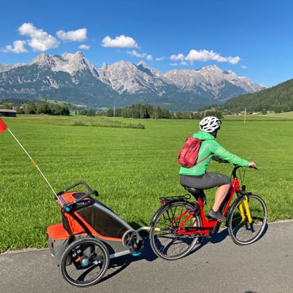 Fietser met kinderaanhanger op pad door groene weide met Steinernes Meer bergketen op achtergrond onder blauwe lucht met witte wolken.