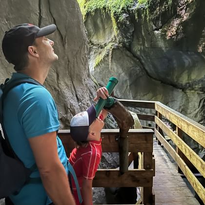 Vader met rugzak en kind met verrekijker op houten loopbrug in Seisenbergklamm kloof, kijkend naar met mos bedekte rotswanden.