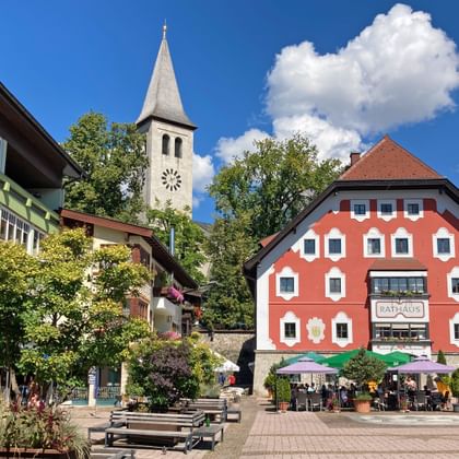 Kleurrijke gebouwen in het centrum van Saalfelden met witte kerktoren, café-terras en bergen op de achtergrond onder blauwe lucht.