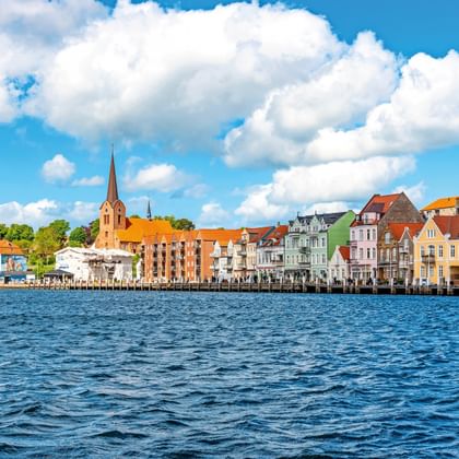 Waterkant van Sønderborg haven met kleurrijke historische gebouwen en kerktoren onder blauwe lucht met witte wolken.