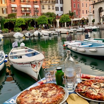 Twee pizza's op borden met drankjes aan watertafel met uitzicht op de haven van Desenzano met kleurrijke boten en historische gebouwen.