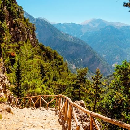Rotsachtig bergwandelpad met houten leuningen door bebost terrein op Kreta, uitkijkend over diepe valleien en verre bergtoppen.