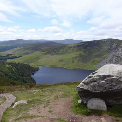 Panoramisch uitzicht vanaf de Wicklow Way op een donker bergmeer omringd door groene heuvels en rotsformaties onder een bewolkte hemel.