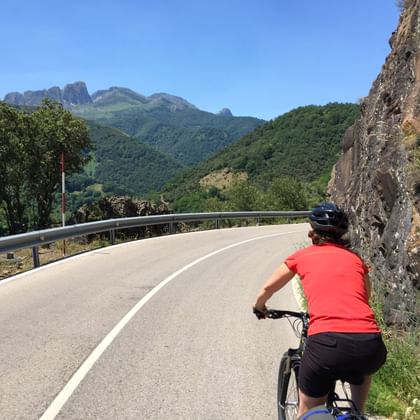 Fietser in rood shirt en helm rijdt langs een kronkelende bergweg in Asturië, Spanje, met rotsachtige kliffen en beboste bergen op de achtergrond.