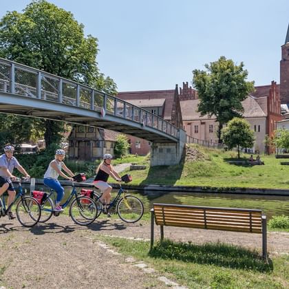 Drie fietsers met fietsen pauzeren bij een rivierpad nabij een moderne brug. Een rode bakstenen kerktoren domineert de achtergrond.