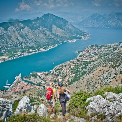 Twee wandelaars met rugzakken op rotsachtig bergpad met uitzicht op de turquoise baai van Kotor met kustplaats en bergen in Montenegro.