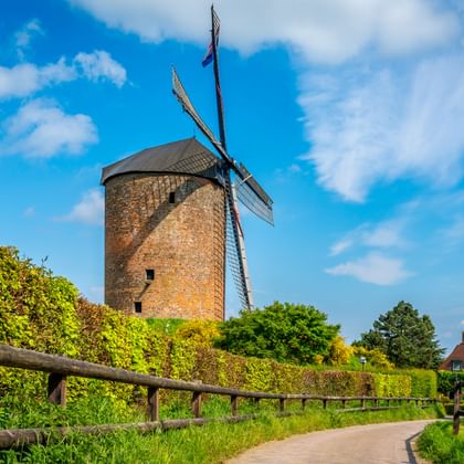 Historische stenen korenmolen met witte wieken onder blauwe lucht in Overijssel. Een houten hek langs een pad naar de molen, omringd door bomen.