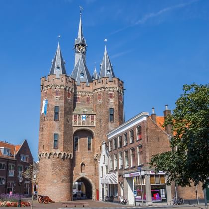 Middeleeuwse bakstenen stadspoort met drie torenspitsen in Zwolle, Overijssel. De rode Sassenpoort staat op een plein met historische gebouwen en bomen.