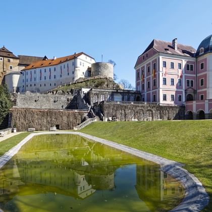 Bečov nad Teplou toont een middeleeuws kasteel op een heuvel en een roze barok paleis met groene koepel. Een vijver op de voorgrond.
