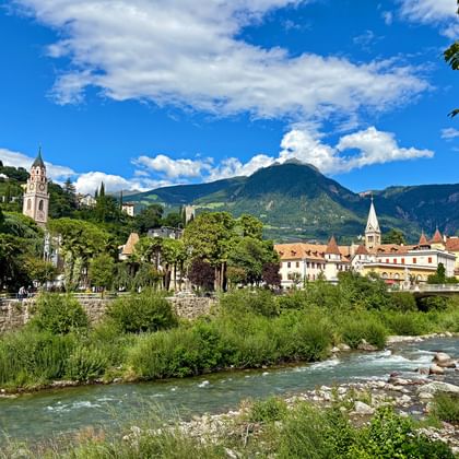 Zicht op Merano met een stromende rivier op de voorgrond, historische gebouwen en kerktorens langs de promenade, omringd door groene bergen.