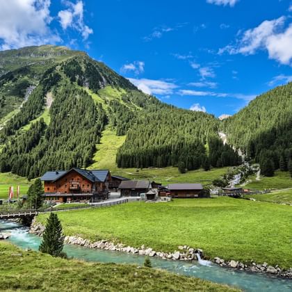 Alpenhutten van Krimmler Tauernhaus in groene vallei met turquoise beek, omringd door beboste bergen onder blauwe lucht in Hohe Tauern.