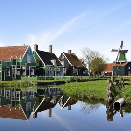Historische groene houten huizen en windmolen in Zaanse Schans weerspiegeld in kalm kanaalwater onder blauwe hemel met witte wolken.