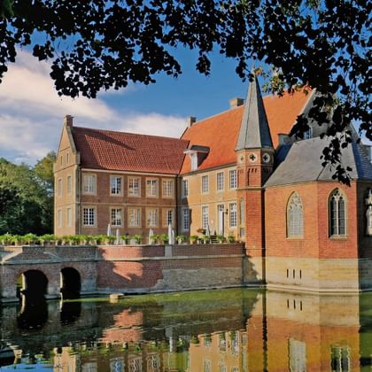 Burg Hülshoff kasteel in Münsterland omgeven door water met stenen brug, rode bakstenen gebouwen en toren weerspiegeld in gracht.