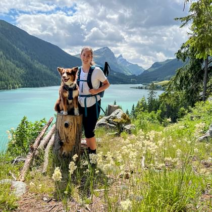 Wandelaar met rugzak en wandelstokken poseert met haar hond op een boomstam met uitzicht op het turquoise Sufnersee omringd door bergen.