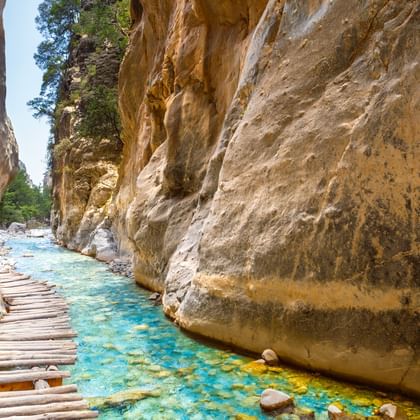 Houten looppad door de Samaria-kloof op Kreta met turquoise beek tussen hoge kalkstenen kliffen en groene vegetatie erboven.