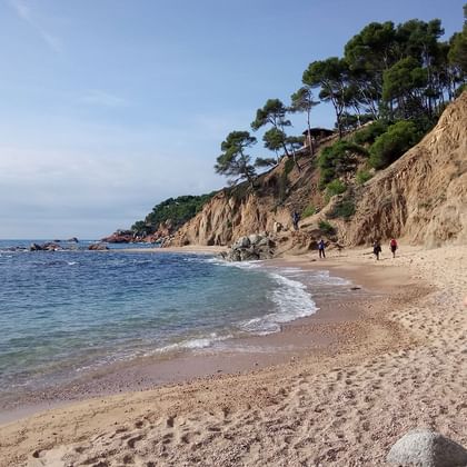 Vrouwelijke wandelaar met rugzak loopt op zandstrand langs Costa Brava kustlijn met dennenbedekte kliffen en helder blauw water.