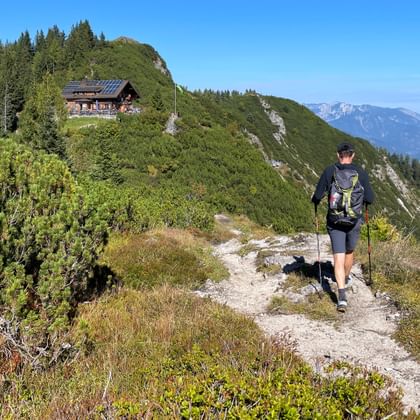 Wandelaar met rugzak op bergpad naar Goiserer Hütte, houten berghut op beboste kam met bergtoppen op de achtergrond.