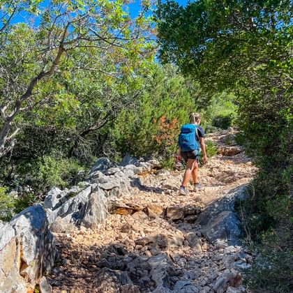 Vrouwelijke wandelaar met blauwe rugzak op rotsachtig gravelpad door mediterraan bos met dichte groene vegetatie en blauwe lucht.