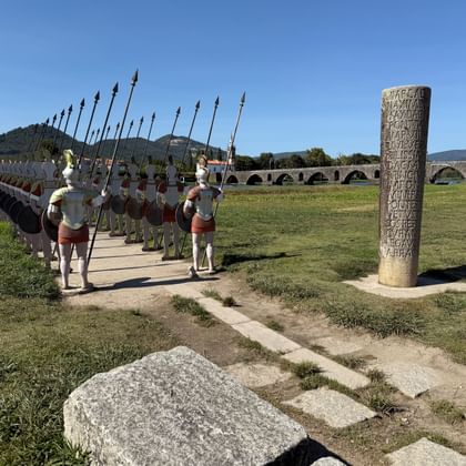 Rij Romeinse soldatenbeelden met speren naast een stenen zuil, met historische boogbrug over Rio Lima op de achtergrond.