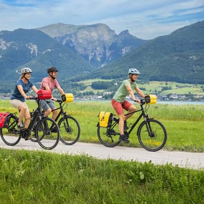Drie fietsers met fietstassen op een pad langs het Wolfgangsee in Salzkammergut, met bergen en een dorp op de achtergrond.