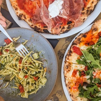Twee pizza's met prosciutto, mozzarella en rucola, en een pastagerecht met pesto op een houten tafel in Piëmont-Ligurië.